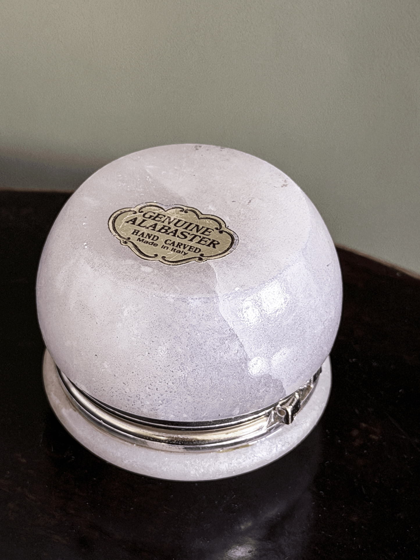 A round, hand-carved alabaster keepsake box with a silver-tone metal band and lid, featuring a gold-colored 'Genuine Alabaster Hand Carved' label on bottom, resting on a dark wooden surface.