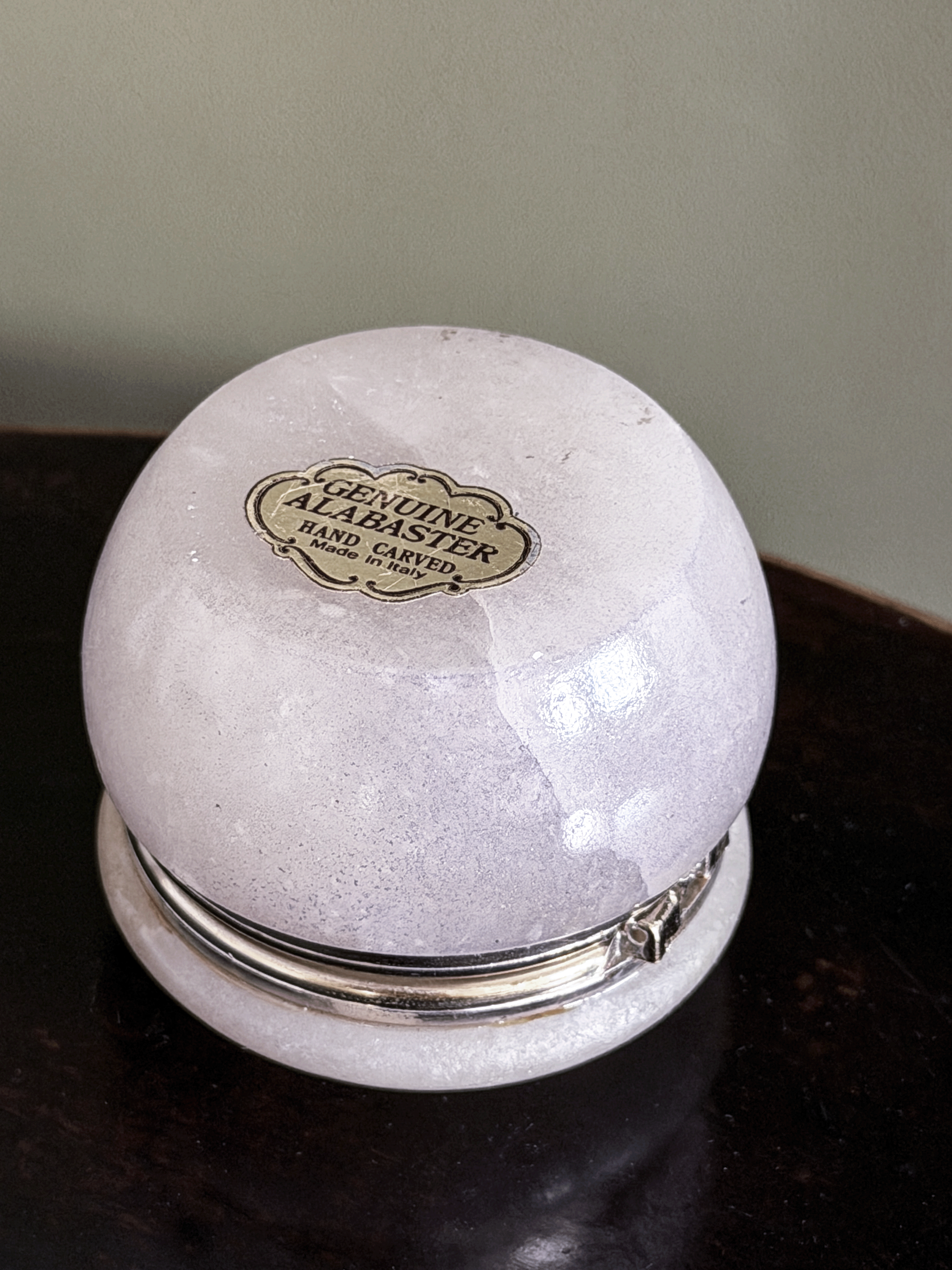 A round, hand-carved alabaster keepsake box with a silver-tone metal band and lid, featuring a gold-colored 'Genuine Alabaster Hand Carved' label on bottom, resting on a dark wooden surface.