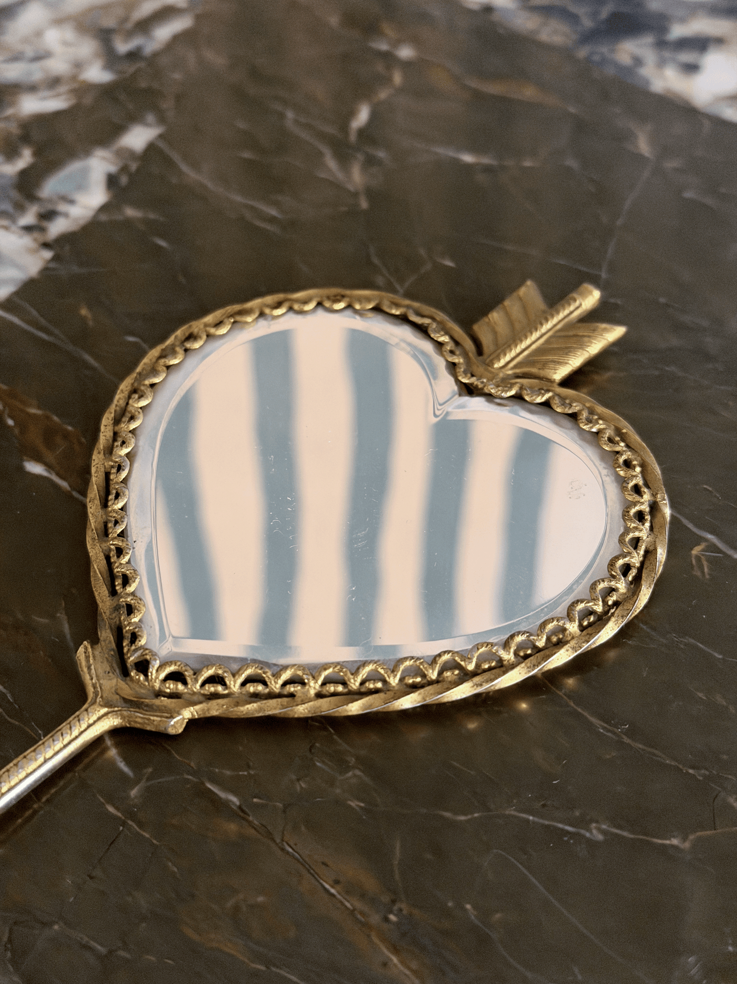 A heart-shaped hand mirror with a gold-toned arrowhead frame and decorative scalloped border, resting on a dark marble surface.