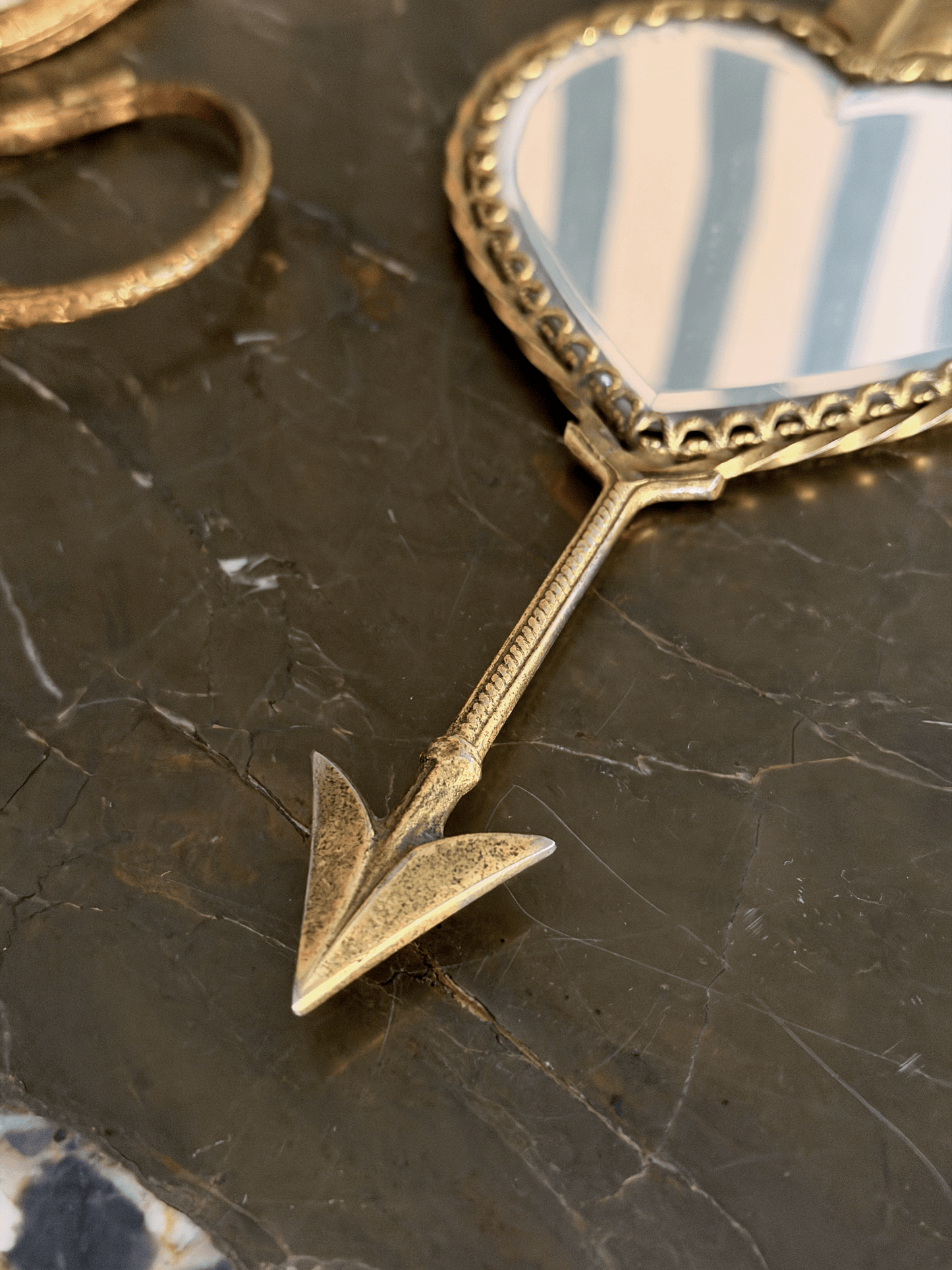 A close-up of a gold-toned decorative mirror with a heart-shaped reflective surface and an arrow-shaped handle, resting on a dark marble surface.