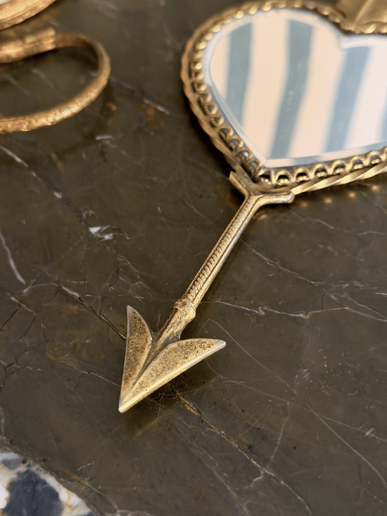 A close-up of a gold-toned decorative mirror with a heart-shaped reflective surface and an arrow-shaped handle, resting on a dark marble surface.