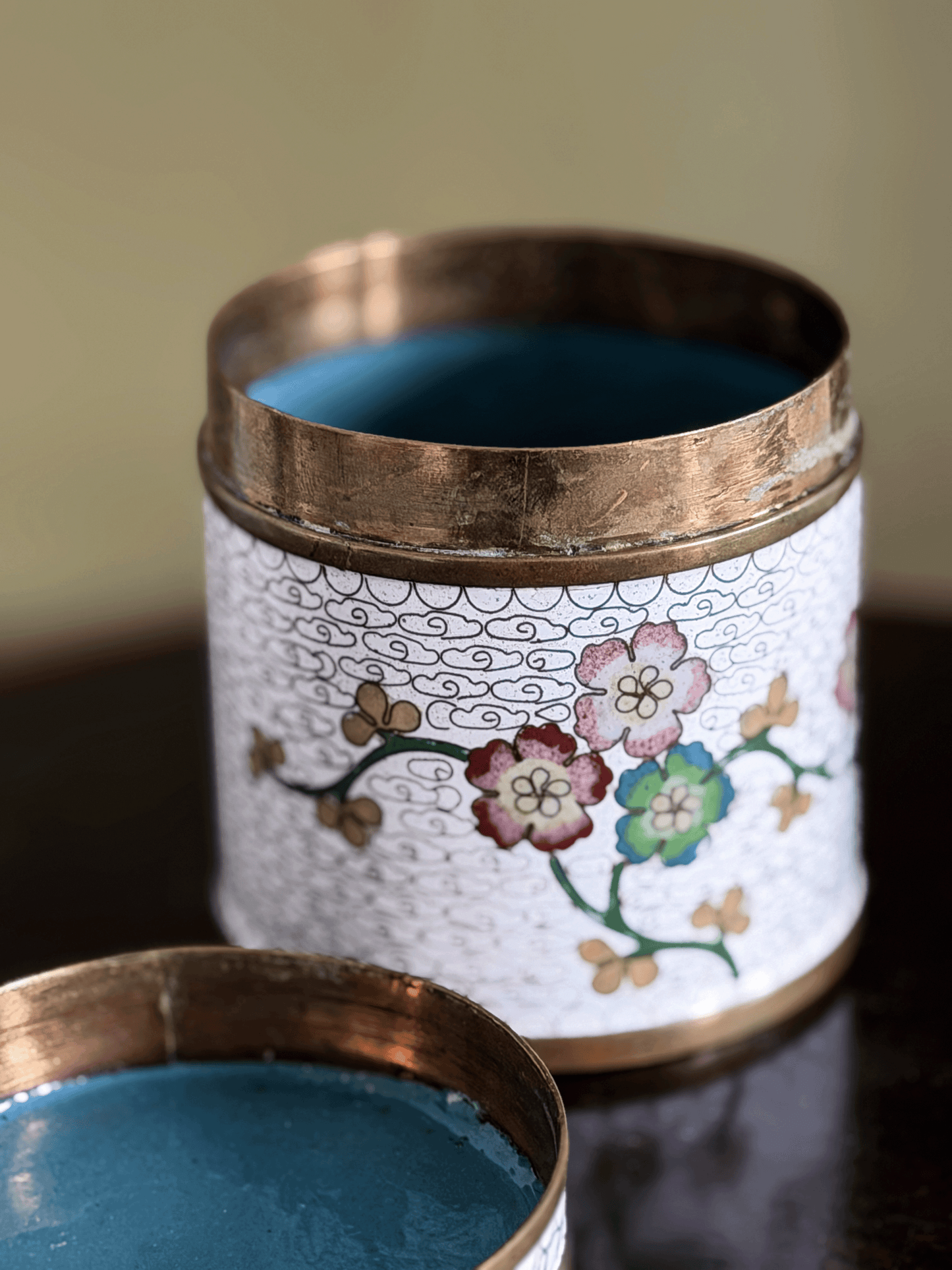 A close-up of a decorative cylindrical tin with a white floral enamel-style body, copper-toned rim, and blue interior, shown open with the lid partially visible in the foreground.