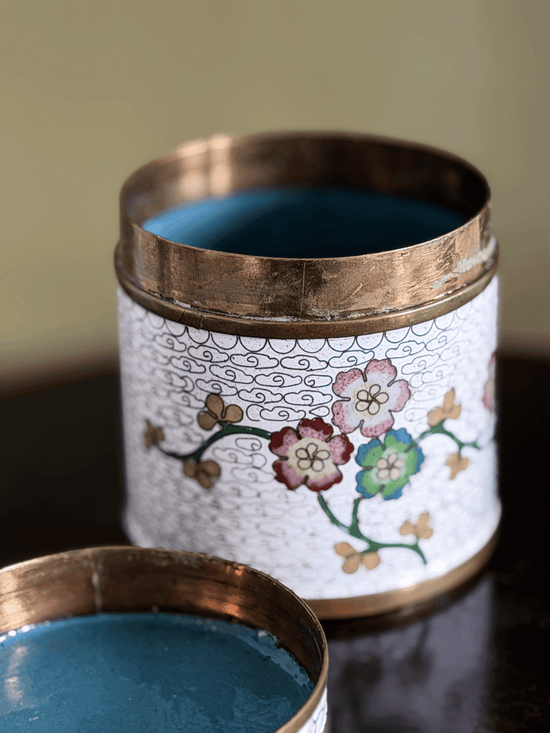 A close-up of a decorative cylindrical tin with a white floral enamel-style body, copper-toned rim, and blue interior, shown open with the lid partially visible in the foreground.