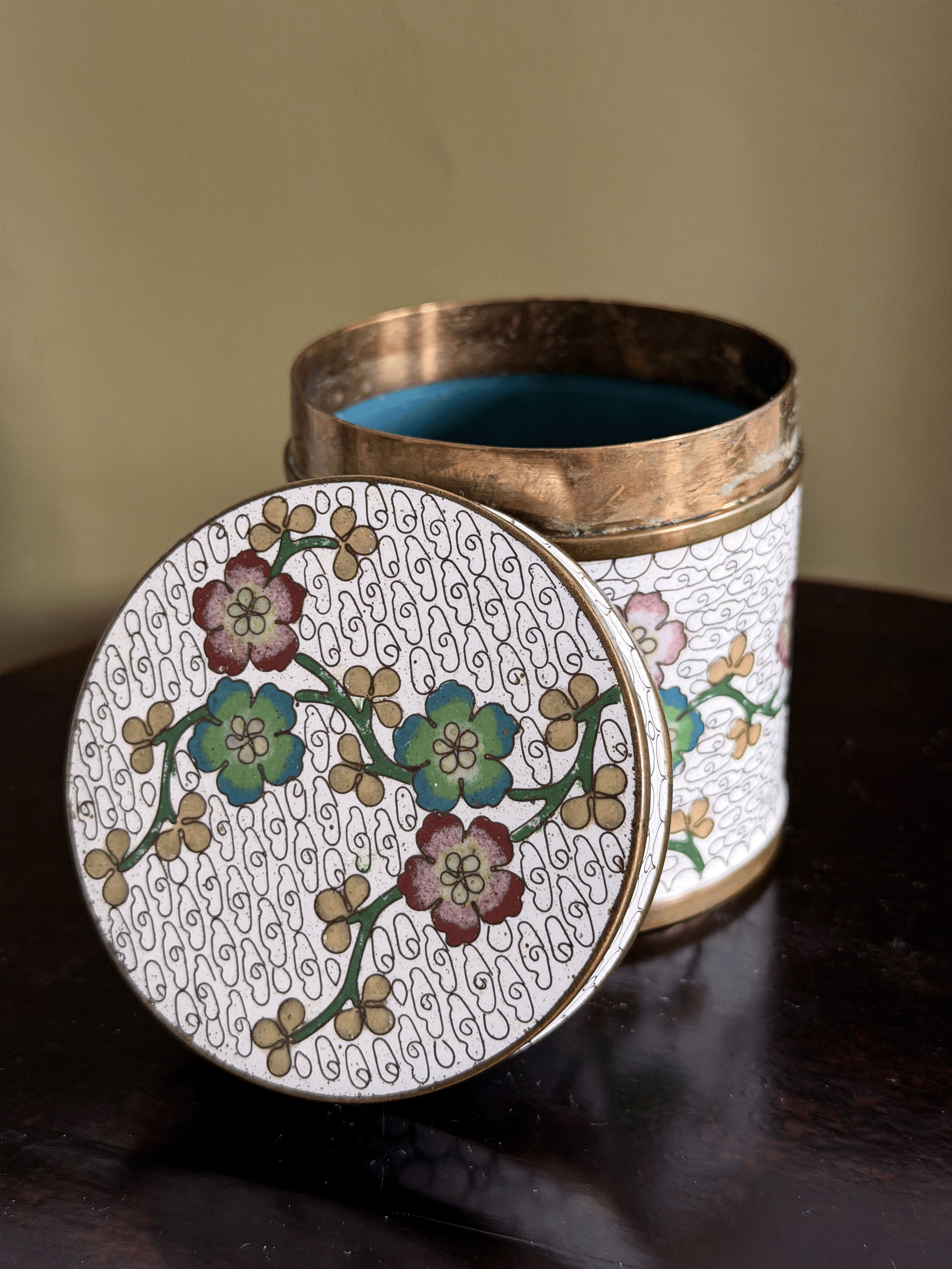 A round decorative metal tin with its lid leaning against it, featuring a white enamel body with a floral pattern, gold-toned scrollwork, and a brass-colored rim, set on a dark wooden surface.