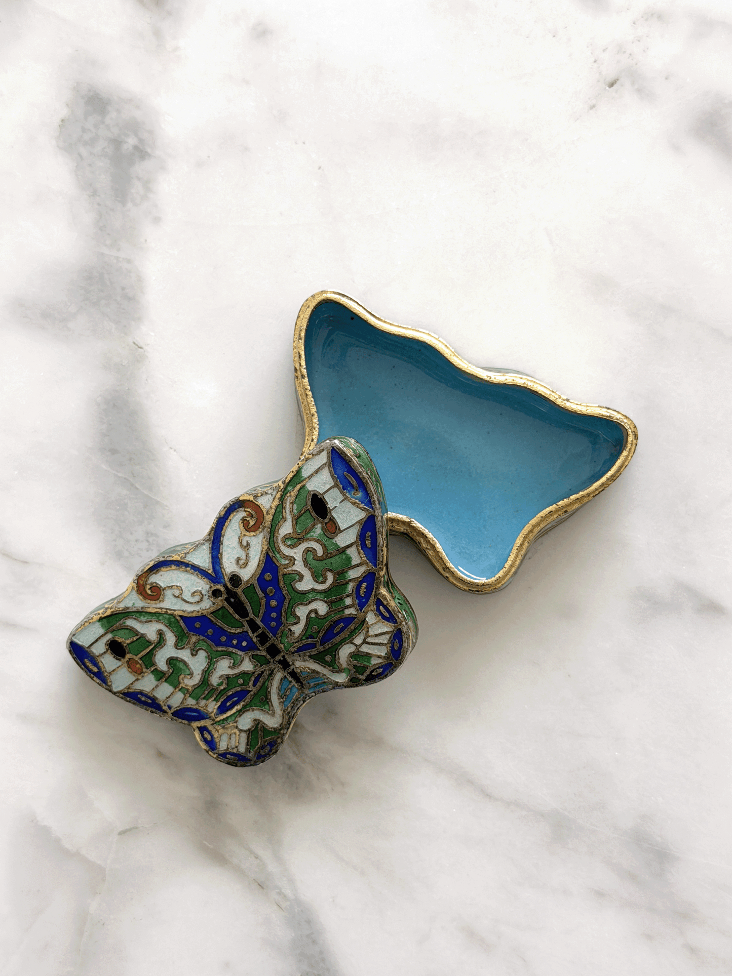A decorative butterfly-shaped trinket box with a light blue enamel interior and gold outline, shown open on a white marble surface. The exterior features a multicolor enamel butterfly design with green, blue, white, and gold details.