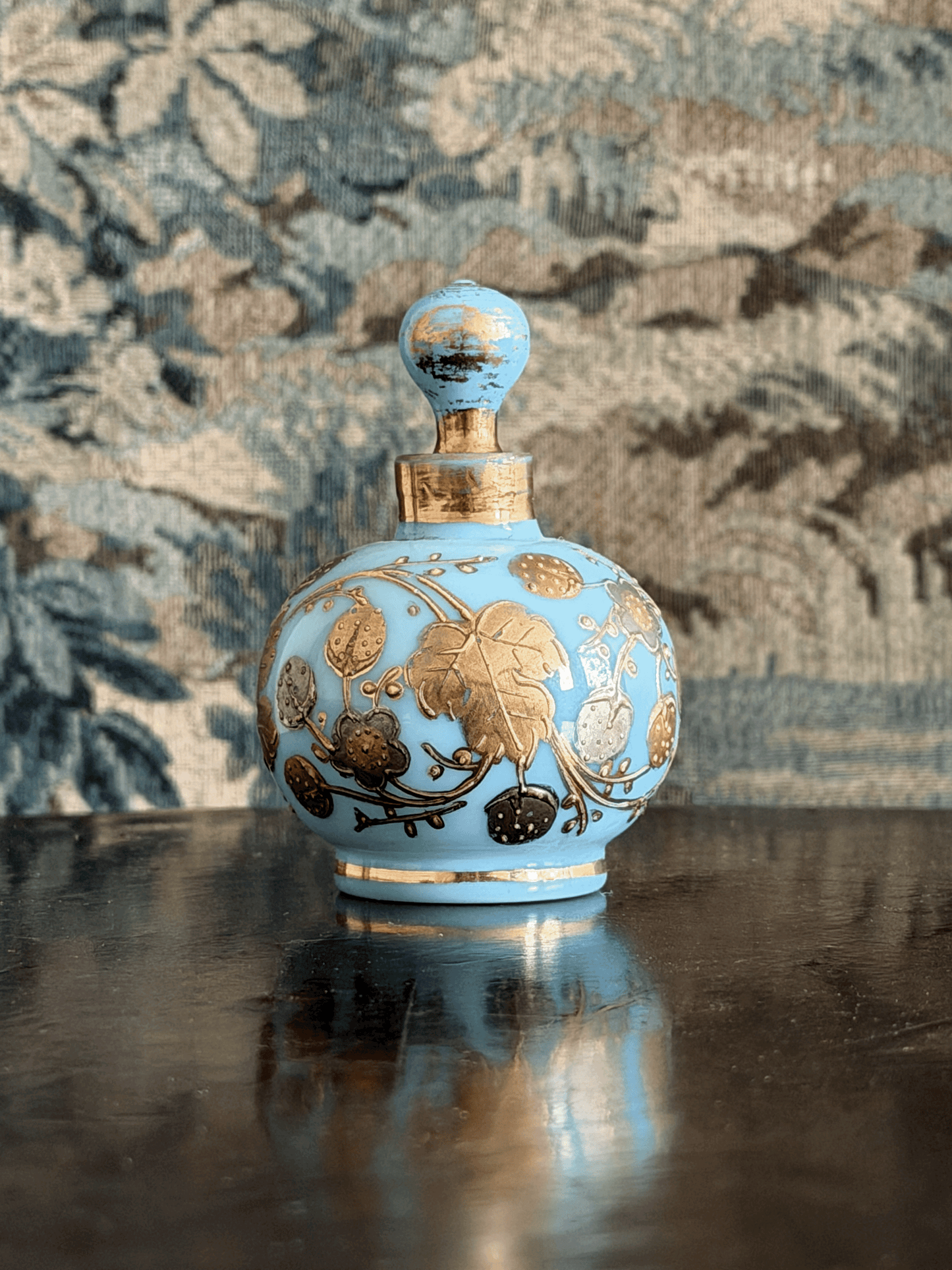 A light blue glass perfume bottle with gold floral detailing and a gold neck ring, displayed on a dark reflective surface against a patterned blue and beige background.