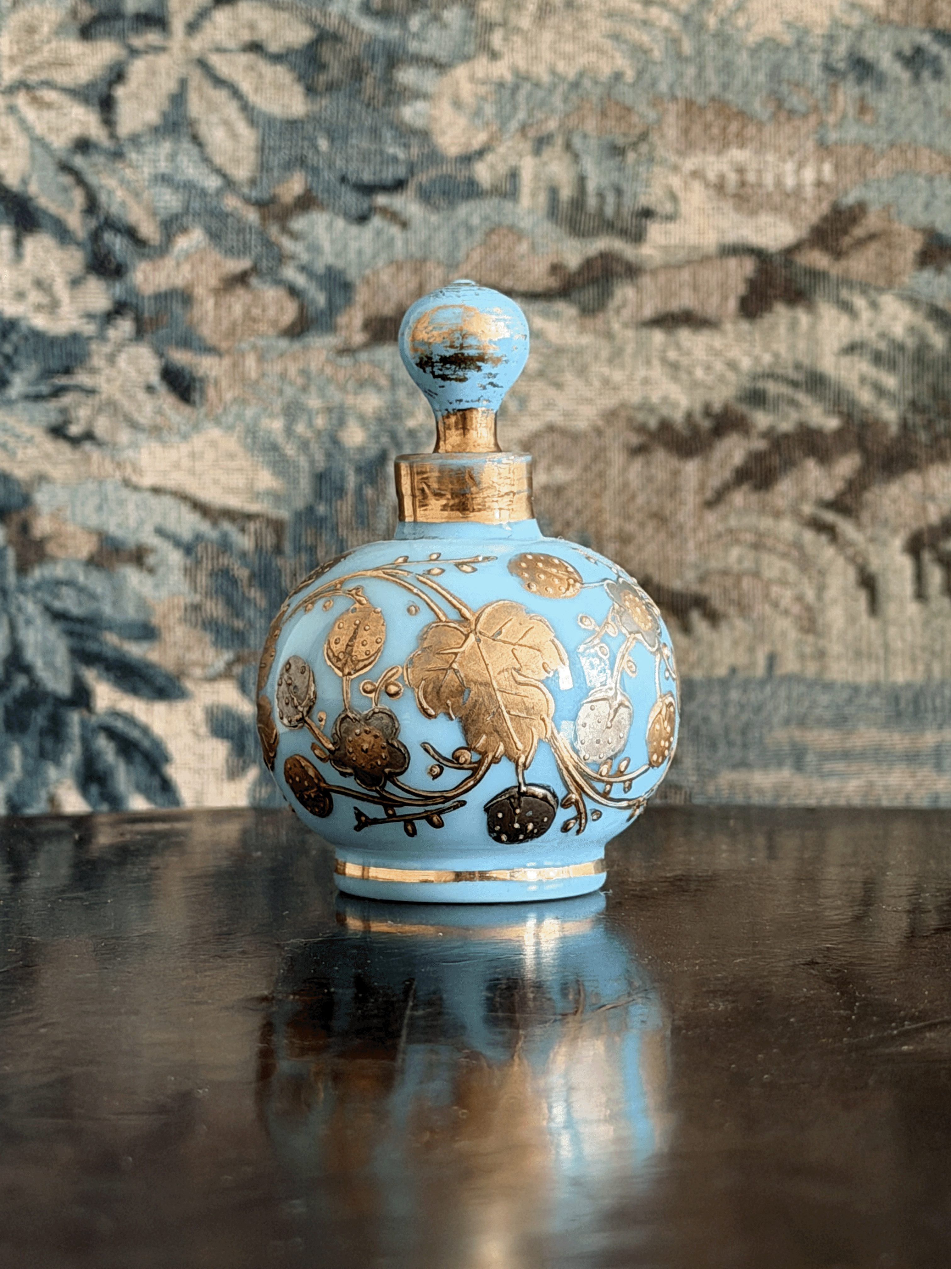 A light blue glass perfume bottle with gold floral detailing and a gold neck ring, displayed on a dark reflective surface against a patterned blue and beige background.