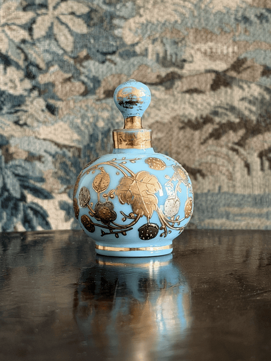 A light blue glass perfume bottle with gold floral detailing and a gold neck ring, displayed on a dark reflective surface against a patterned blue and beige background.