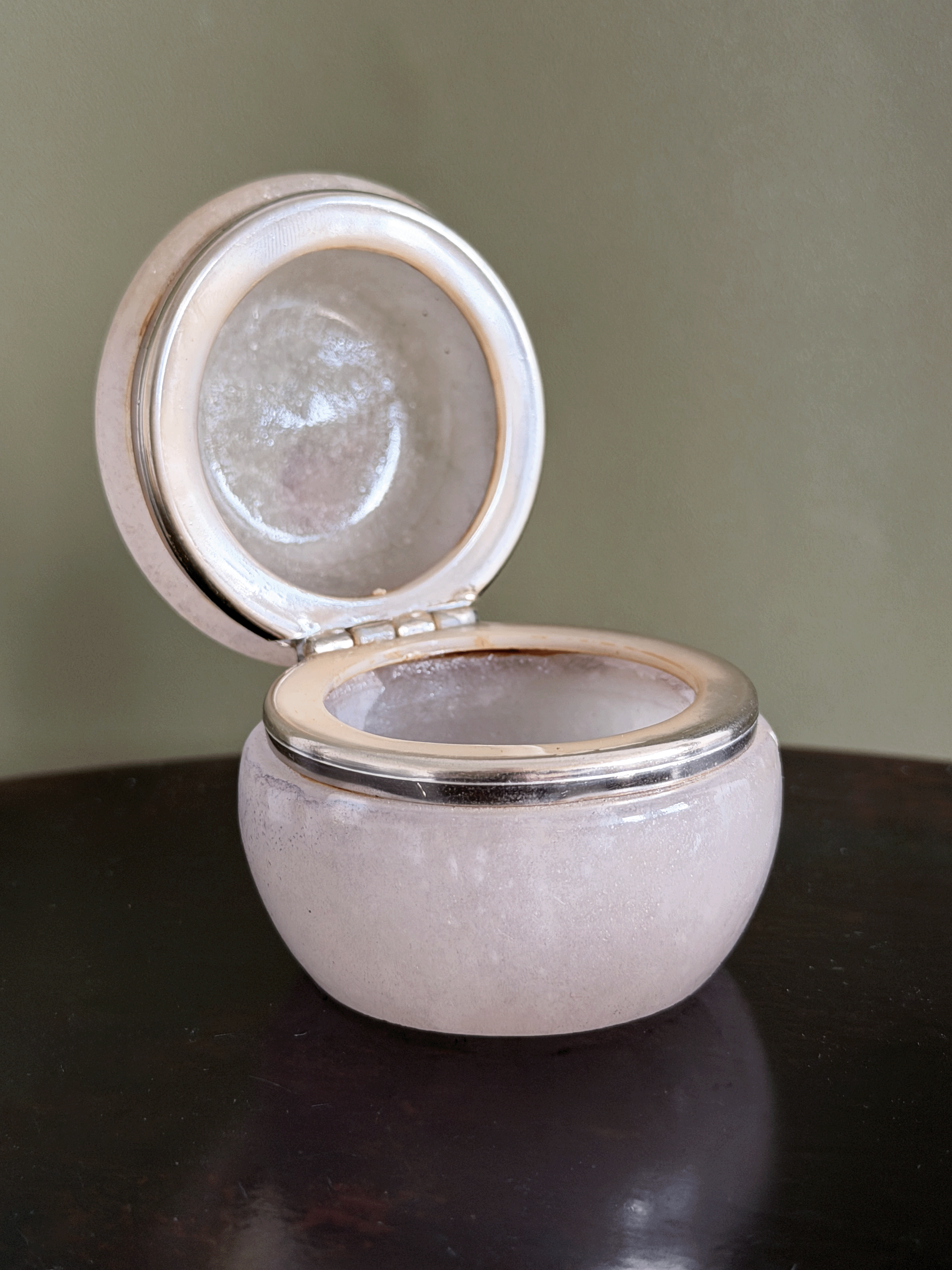A small round alabaster stone keepsake box with a hinged lid, shown open on a dark reflective surface against a muted olive-green background.