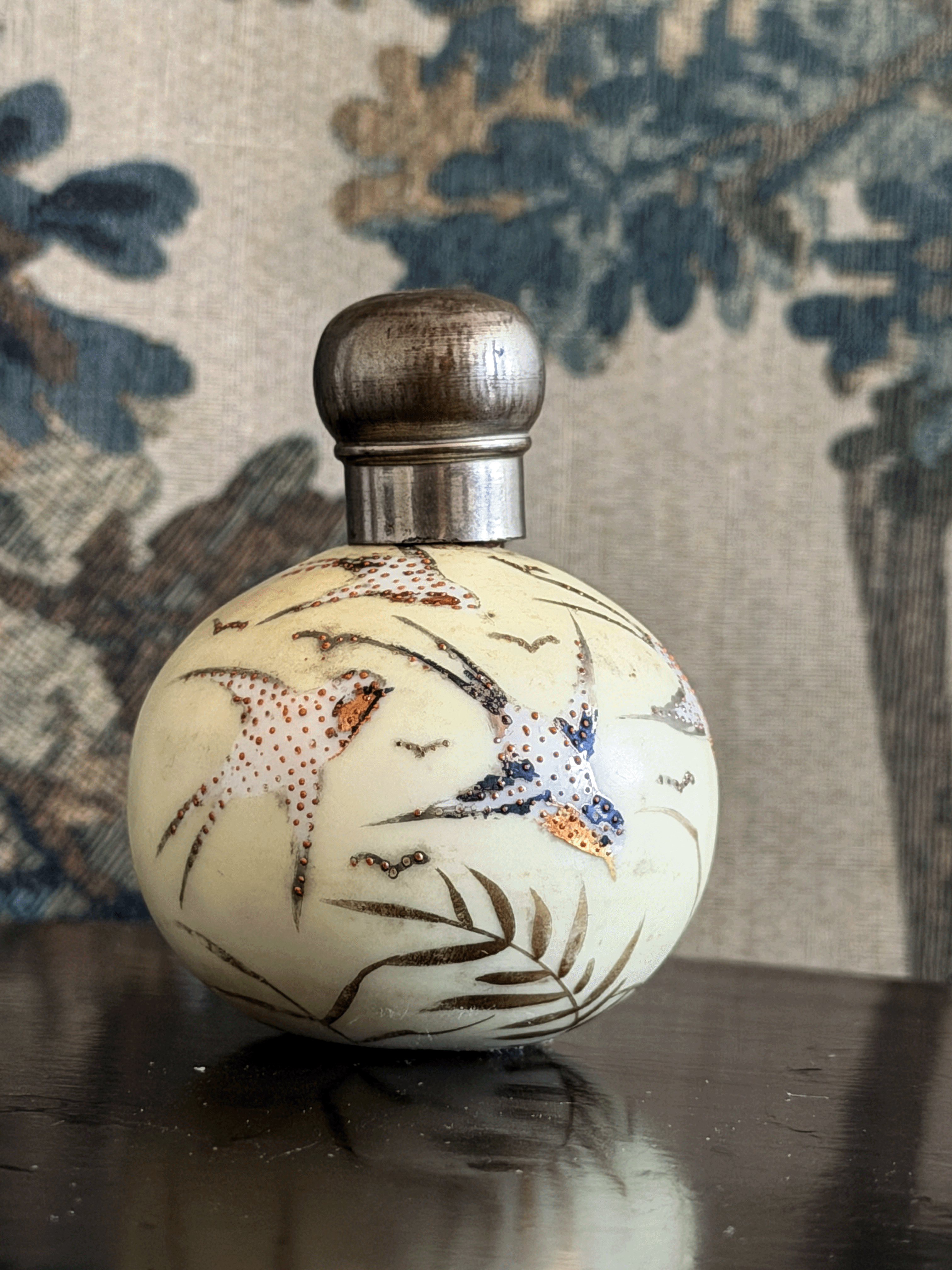 A small round perfume bottle with a silver-toned metal cap, decorated with hand-painted birds and leaves in brown, blue, and orange, set against a patterned fabric background.