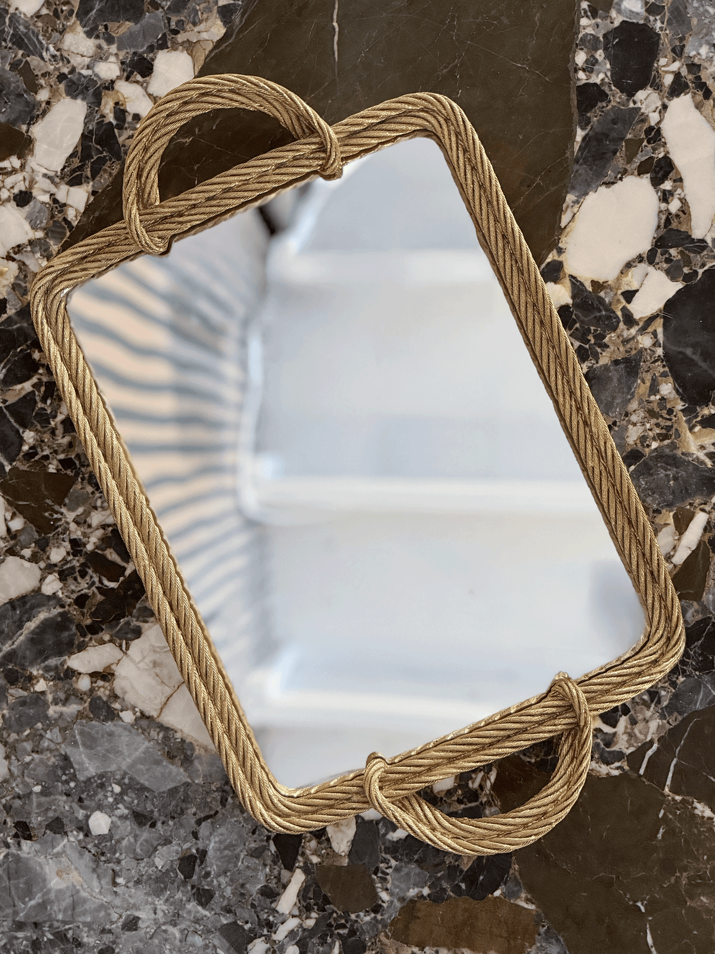 A rectangular mirror with a thick, twisted rope-style frame in a warm gold finish, resting on a dark, polished marble surface.