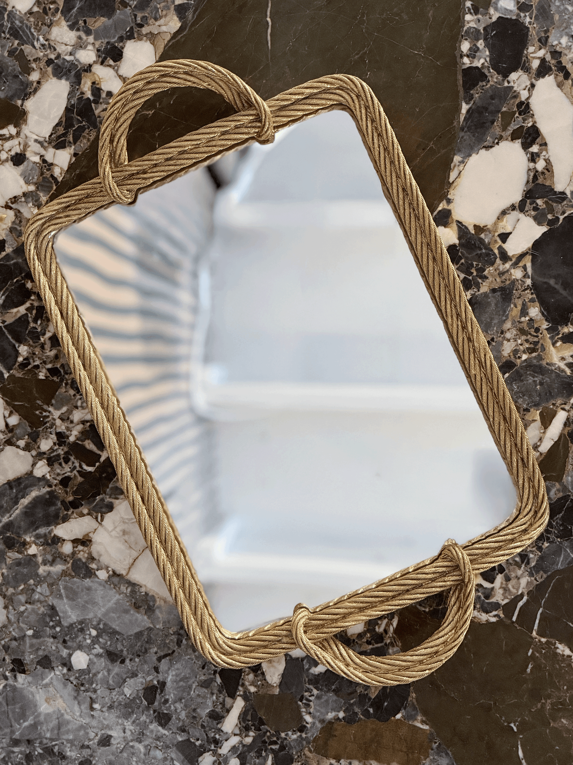 A rectangular mirror with a thick, twisted rope-style frame in a warm gold finish, resting on a dark, polished marble surface.