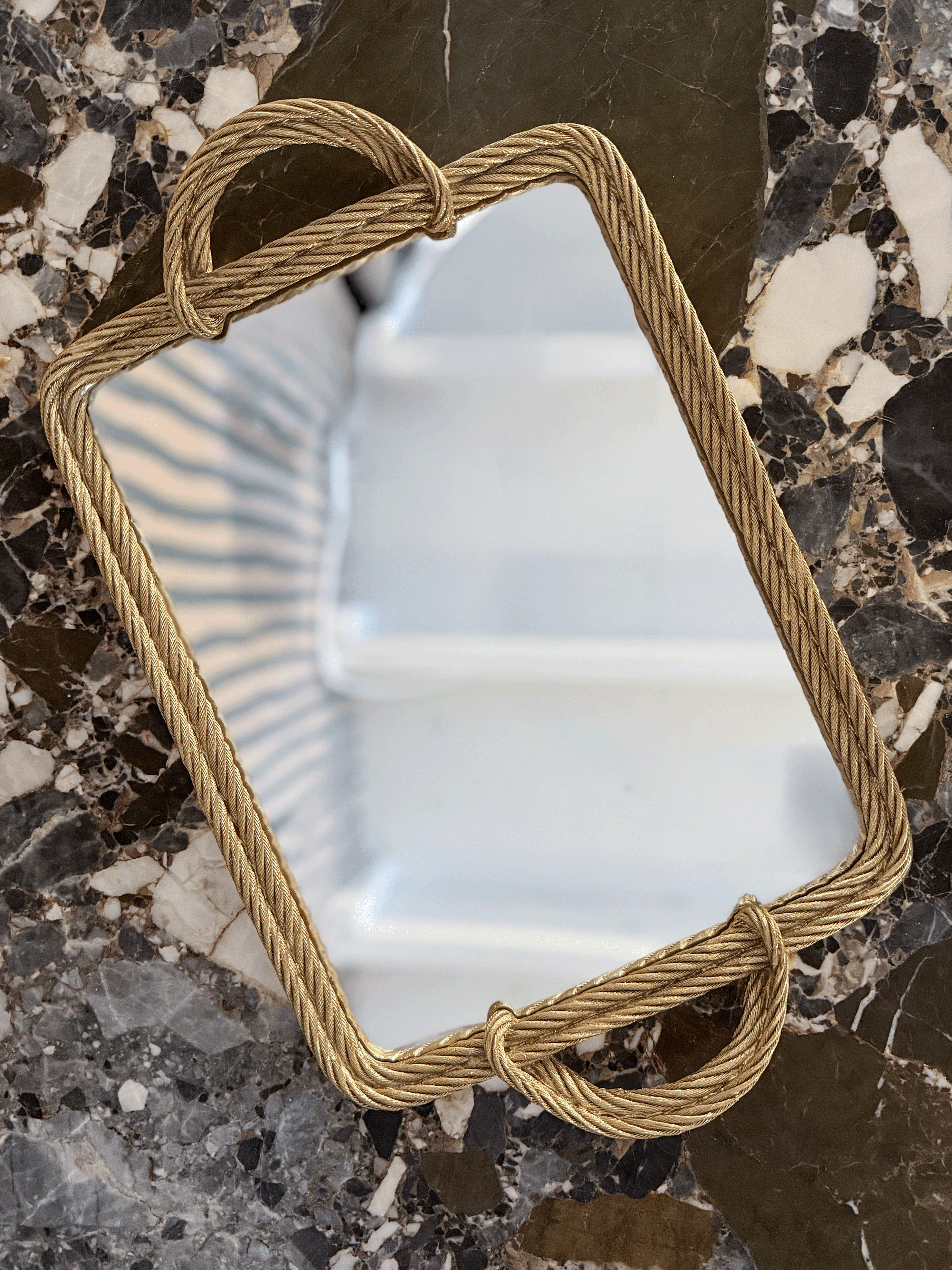 A rectangular mirror with a thick, twisted rope-style frame in a warm gold finish, resting on a dark, polished marble surface.