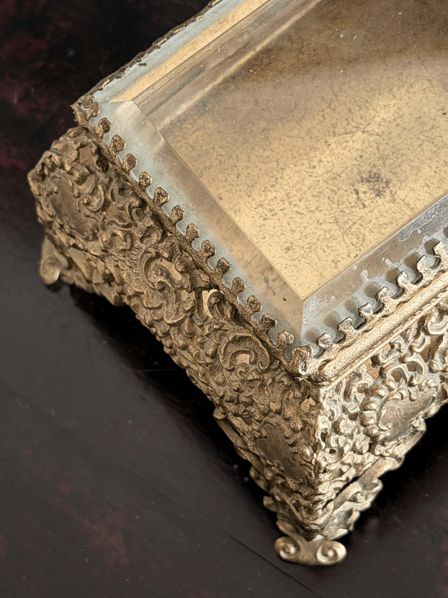 An ornate antique-style gold-toned metal jewelry box with intricate floral scrollwork and a clear glass lid, shown on a dark wooden surface.