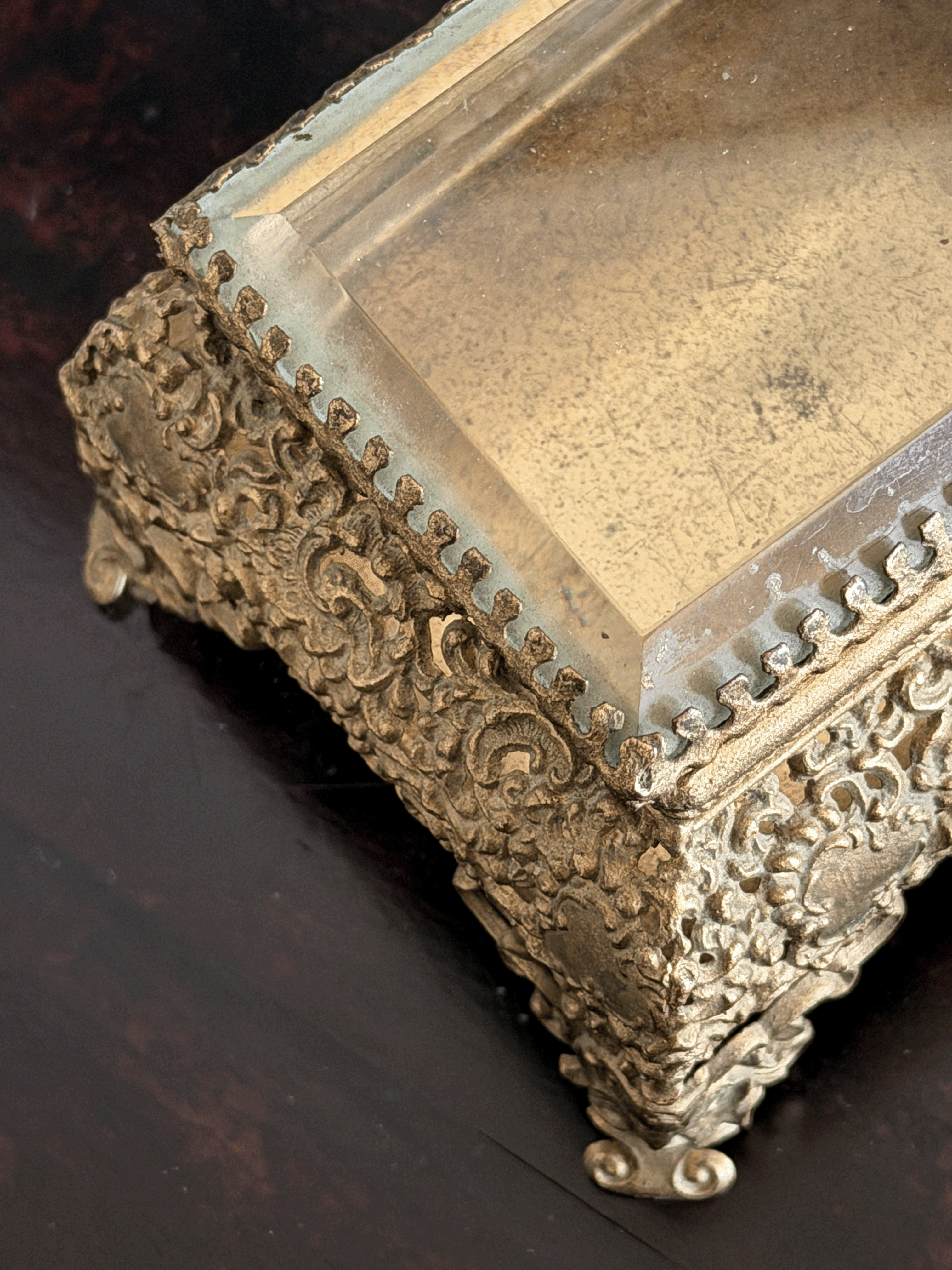 An ornate antique-style gold-toned metal jewelry box with intricate floral scrollwork and a clear glass lid, shown on a dark wooden surface.
