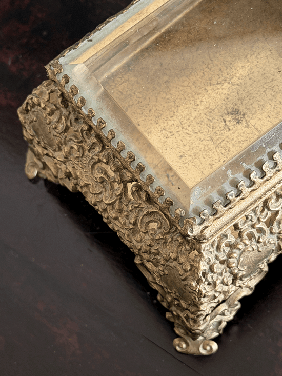 An ornate antique-style gold-toned metal jewelry box with intricate floral scrollwork and a clear glass lid, shown on a dark wooden surface.