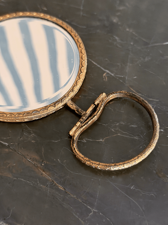 An antique-style oval hand mirror with a distressed gold frame and a matching ring pull handle
