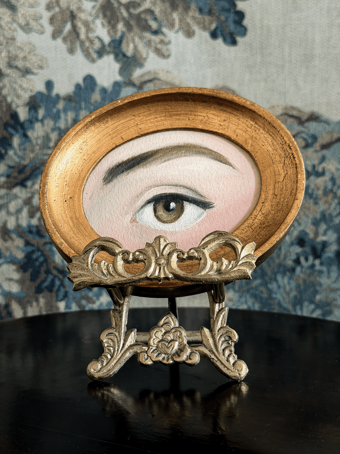A decorative oval art piece featuring a painted human eye, displayed on an ornate gold stand against a blue floral patterned backdrop.