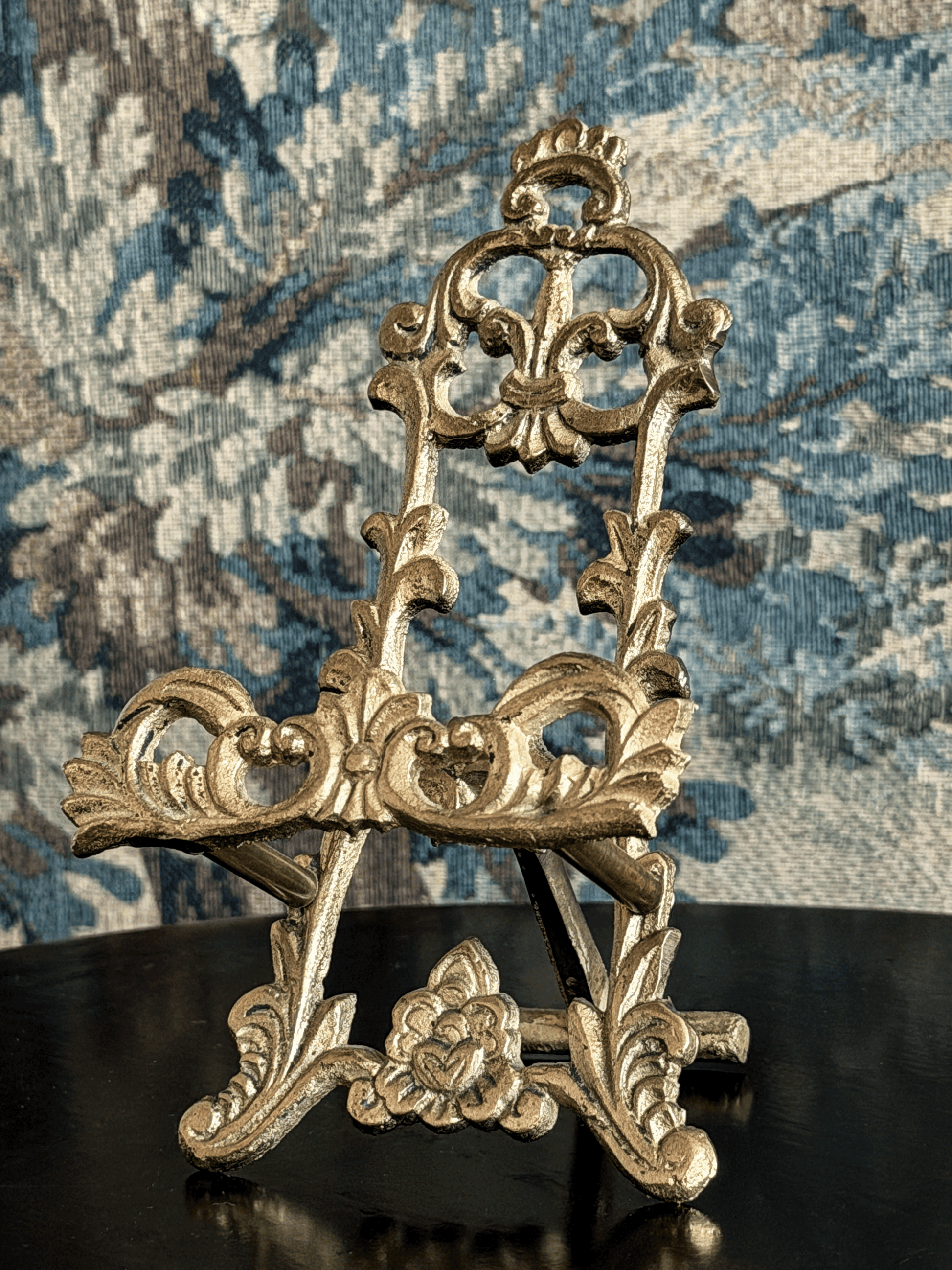 An ornate gold-colored metal miniature display stand with intricate baroque-style scrollwork and floral details, shown on a dark reflective surface against a blue patterned tapestry background.
