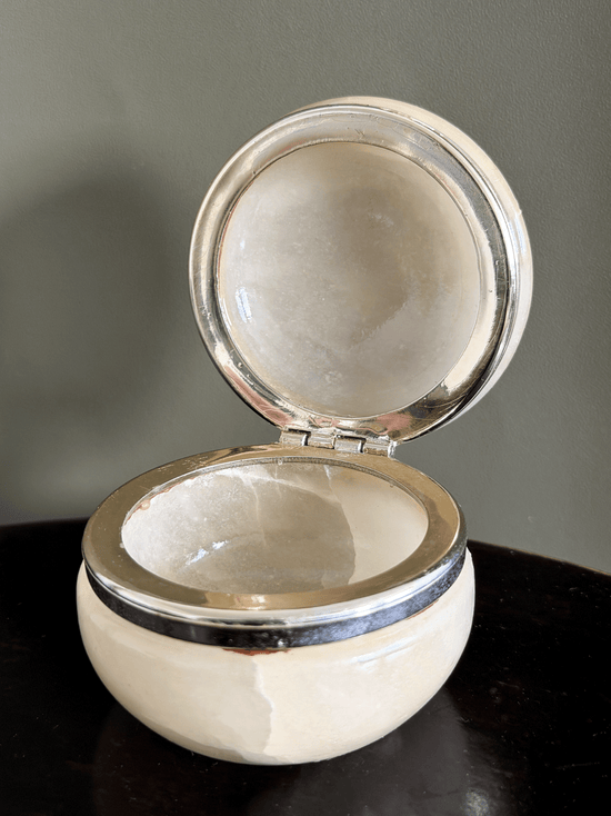 A small round cream-colored jewelry box with a polished silver-tone metal rim and open hinged lid, displayed on a dark surface against a muted gray background.
